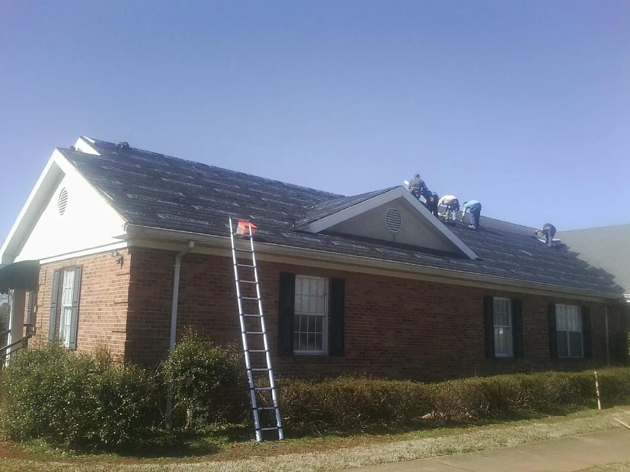 Crew tearing shingles off a brick ranch roof with a ladder set — Emergency Roof Repair work by our crew in Elon