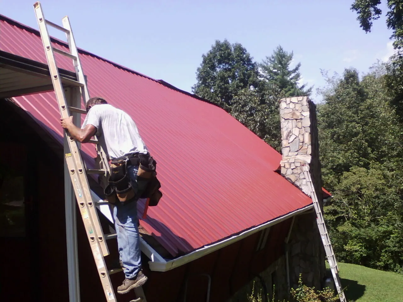 Roofer on a ladder beside a red metal roof and stone chimney — Soffit & Fascia Repair by Roofing Brothers in Elon